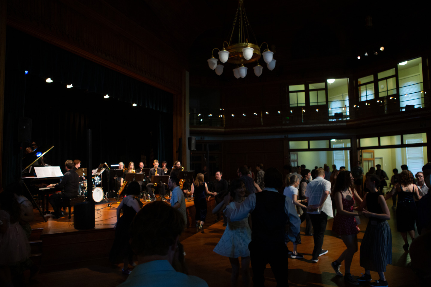 The band on stage while couples swing dance across the ballroom floor beneath a chandelier.